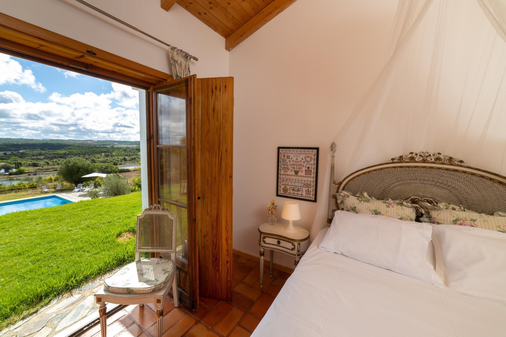 Een slaapkamer met een groot raam, een bed, een nachtkastje en uitzicht op een zwembad en groen.