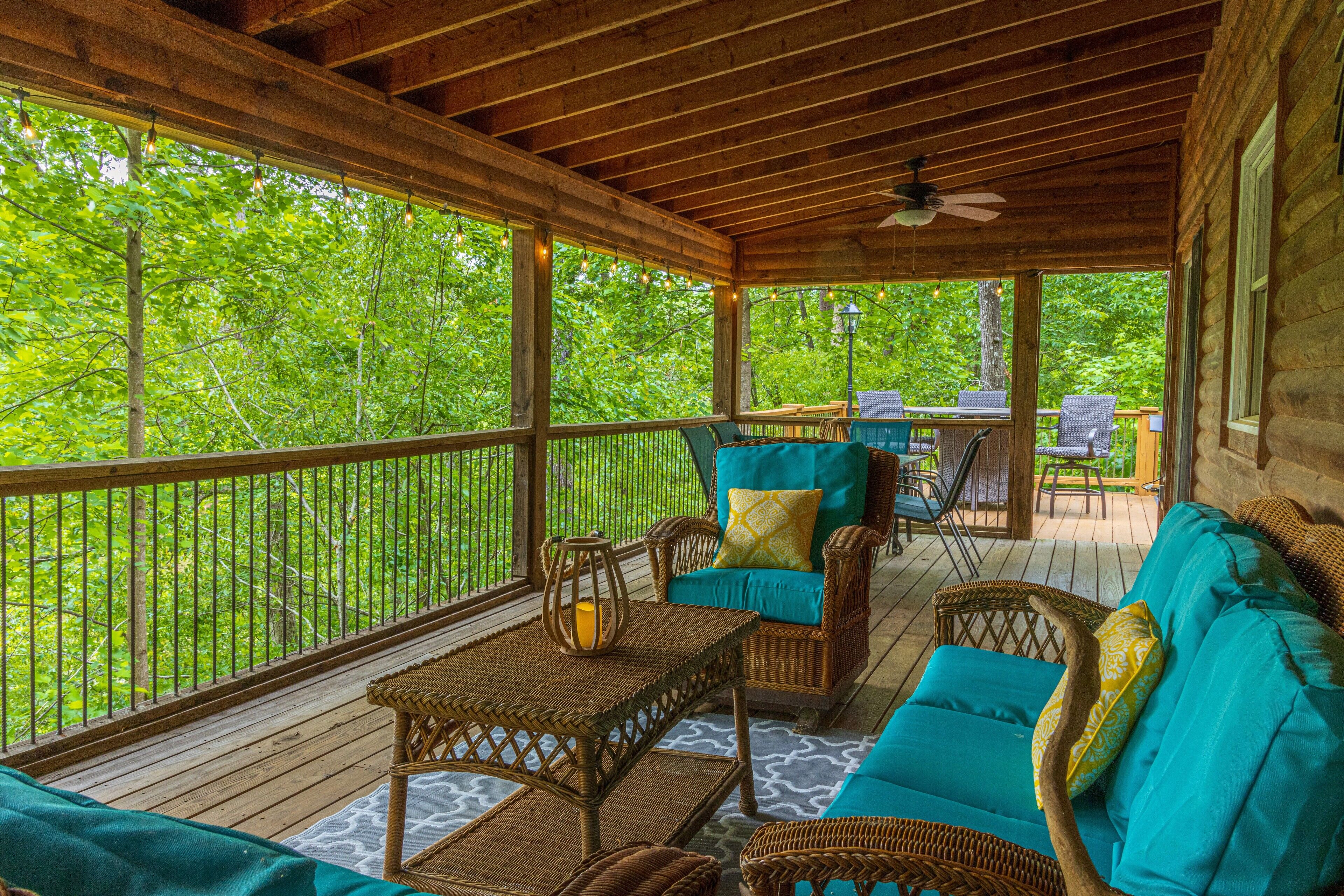 A covered porch with wicker furniture, a table with a lantern, and a view of lush greenery.