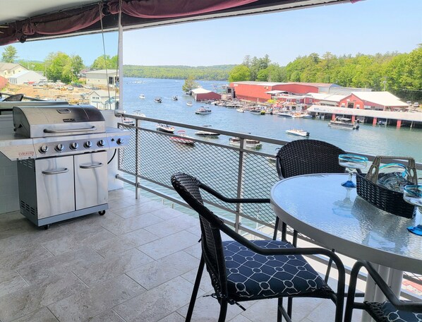 Een balkon met een barbecue, stoelen en een tafel, met uitzicht op een jachthaven met boten en gebouwen.