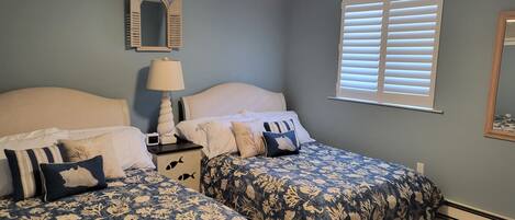 Une chambre avec deux lits, un ventilateur de plafond, une table de chevet avec une lampe, un miroir et une fenêtre avec des stores.