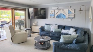Ein modernes Wohnzimmer mit einem blauen Sofa, einem beige farbenen Sessel und einem runden Couchtisch. An der Wand ist ein Flachbildfernseher angebracht, und es sind Wandbilder im maritimen Stil zu sehen.