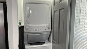 A compact laundry room with a front-loading washer and dryer, a small shelf with a bottle, and a door leading to another room. - Top-Rated Home Near Ball State & IU Hospital- Top Guest Pick!  (Muncie)