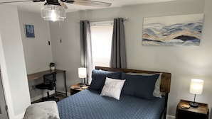 A bedroom with a bed, two bedside tables, a desk, a chair, a ceiling fan, a painting, and a window with curtains. - Top-Rated Home Near Ball State & IU Hospital- Top Guest Pick!  (Muncie)