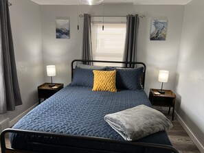 A neatly made bed with a blue quilt, two pillows, and a folded blanket. There are two bedside tables with lamps, a window with blinds, and two wall art pieces. - Top-Rated Home Near Ball State & IU Hospital- Summer Specials (Muncie)