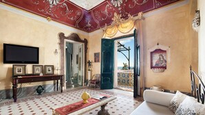 A luxurious room with a marble floor, a large bed with a white headboard, a wooden console table with framed pictures, a television, and a view of a balcony with a tiled floor. - Palazzo Crocione in Positano (Positano)