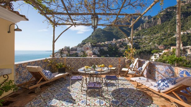 A terrace with a tiled floor, outdoor seating, a table with fruit, and a view of a coastal town. - Palazzo Crocione in Positano (Positano)