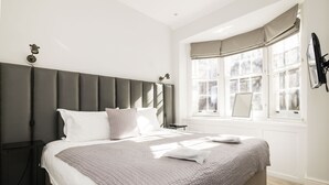 A bedroom with a large bed, a headboard, a nightstand, a window with curtains, and a wall-mounted lamp.