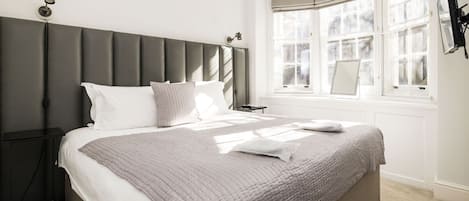 A bedroom with a large bed, a headboard, a nightstand, a window with curtains, and a wall-mounted lamp.