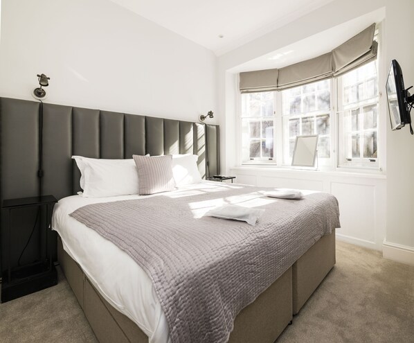 A bedroom with a large bed, a headboard, a nightstand, a window with curtains, and a wall-mounted lamp.