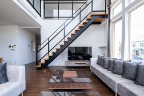 A modern living room with a glass coffee table, a sofa with cushions, a staircase, and a television. - THE BEEHIVE KAMIFURANO (Kamifurano)