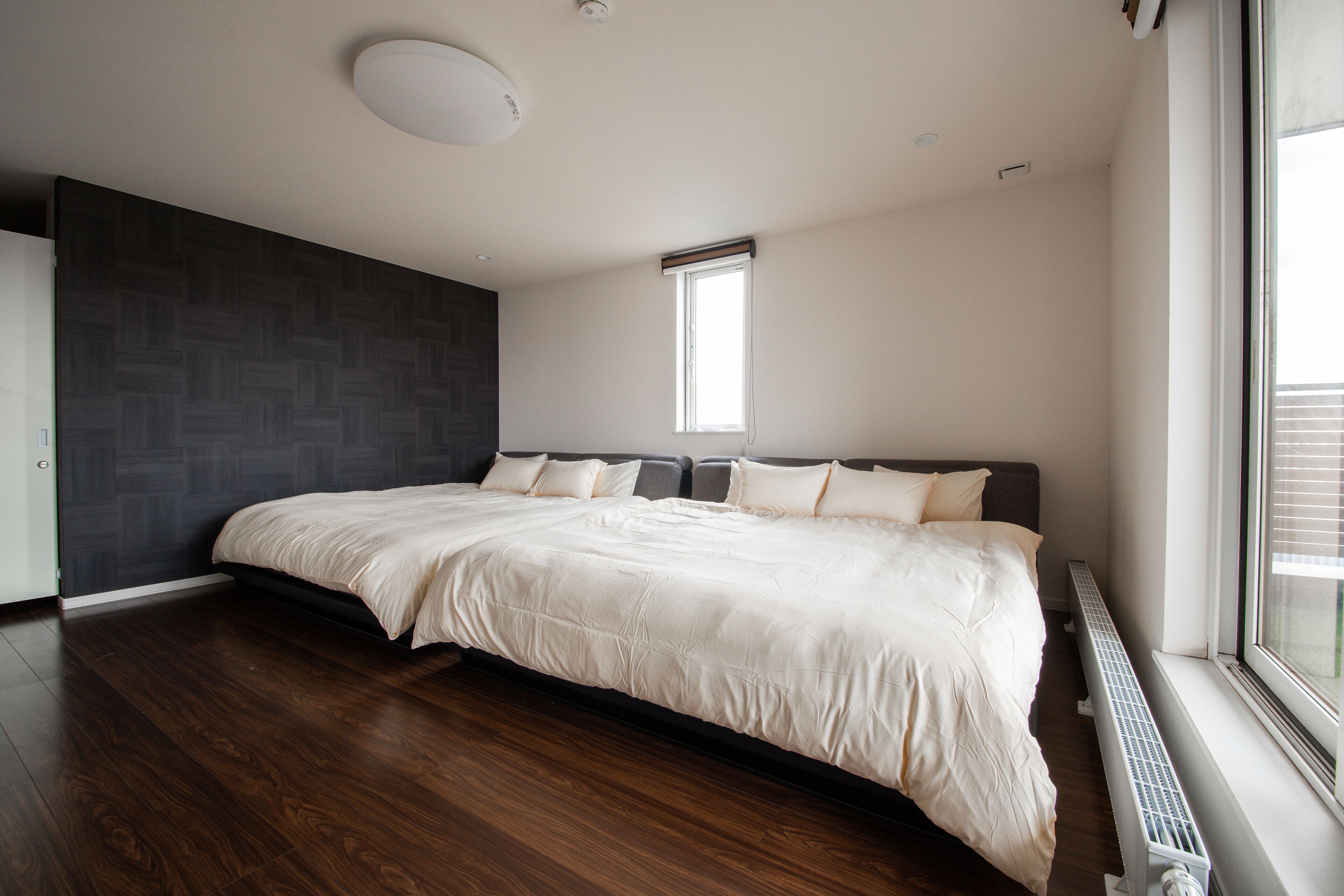 A modern bedroom with a large bed, dark wooden flooring, a built-in wardrobe, and a window providing natural light.