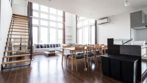 A spacious living area with a wooden staircase, a dining table with chairs, a sofa, and a kitchen area with a sink and countertop. - THE BEEHIVE KAMIFURANO (Kamifurano)