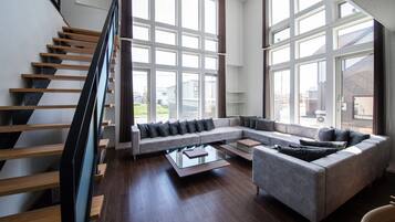 A spacious living room with a wooden staircase, large windows, and a grey sectional sofa.