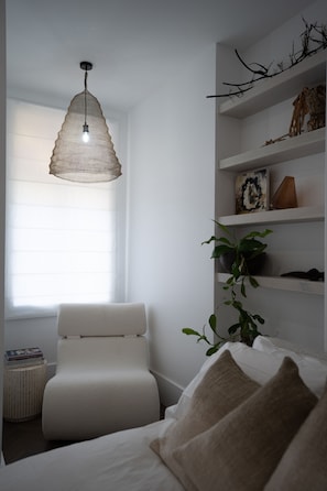 Een slaapkamer met een bed, een witte stoel, een nachtkastje met boeken, een hanglamp en planken met decoratieve objecten.