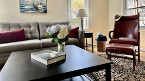 Un salon avec un canapé, une table basse sur laquelle se trouve un livre, un vase rempli de fleurs, une lampe et un fauteuil sur lequel est posé un chapeau.