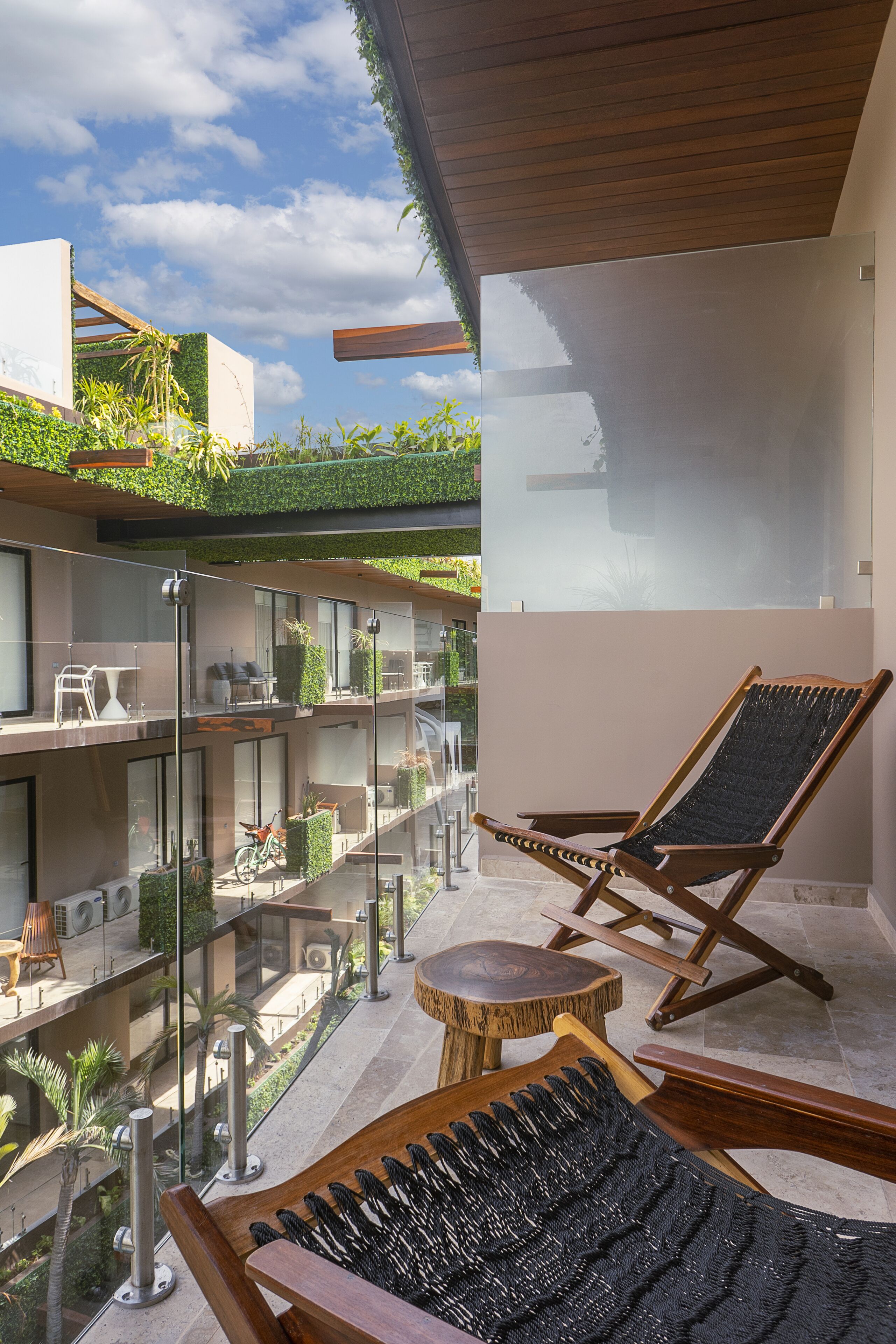A modern outdoor lounge area with a glass balustrade, wooden lounge chairs, and a small round table.