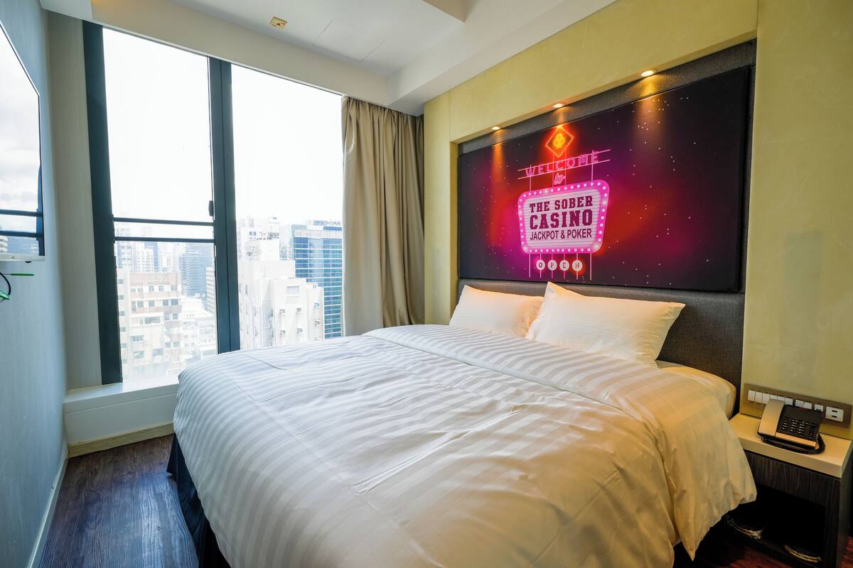A hotel room with a large bed, a city view, and a neon sign advertising The Sofe Casino.