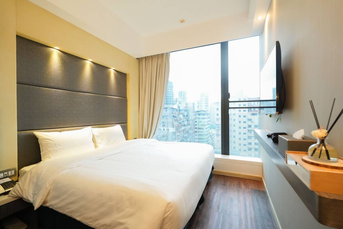 A modern hotel room with a large bed, a flat-screen TV mounted on the wall, a wooden nightstand, and a view of the city through a large window.