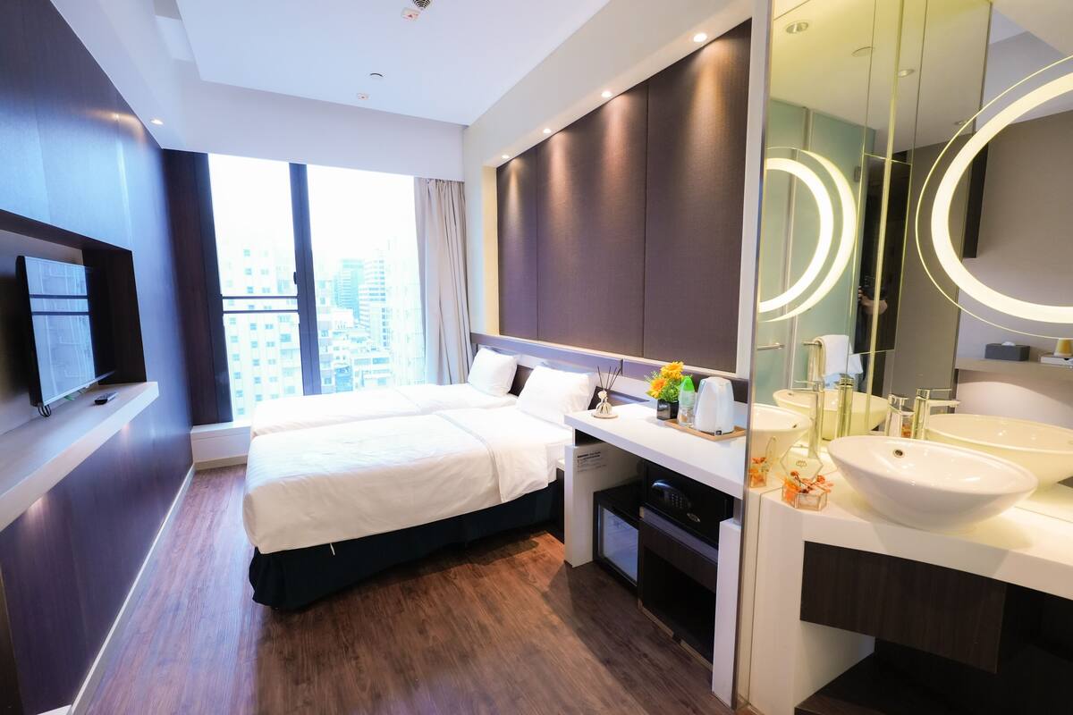 A modern hotel room with a large bed, a vanity with a large mirror, and a view of the city through large windows.