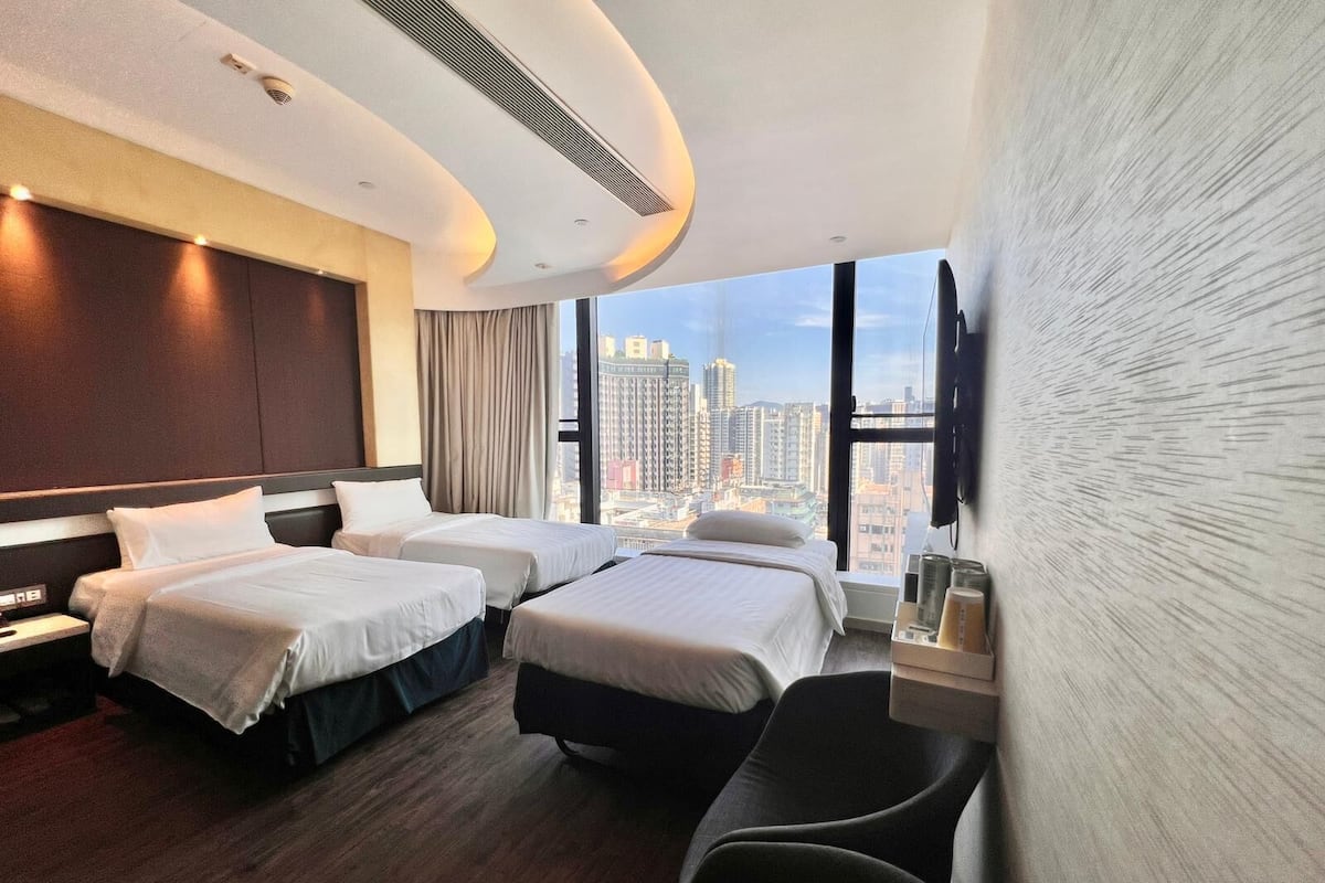 A hotel room with two beds, a desk, a chair, and a large window offering a city view.