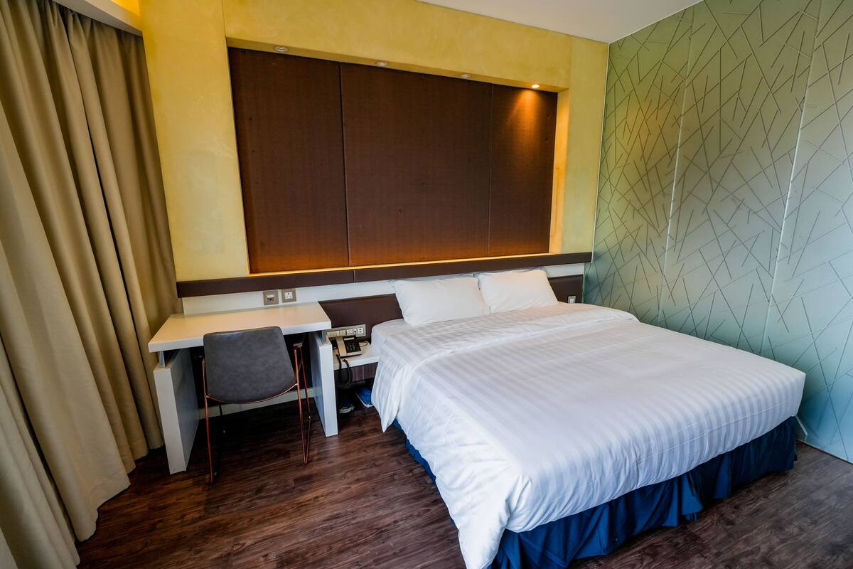 A hotel room with a large bed, a desk with a chair, and a wardrobe.