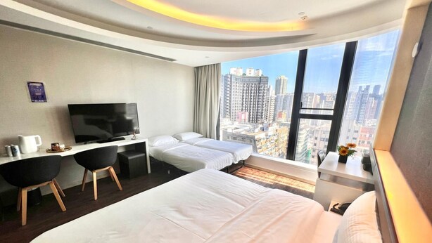 A hotel room with a large bed, a desk with a TV, and a view of the city. - Epoch Hotel (Kowloon)