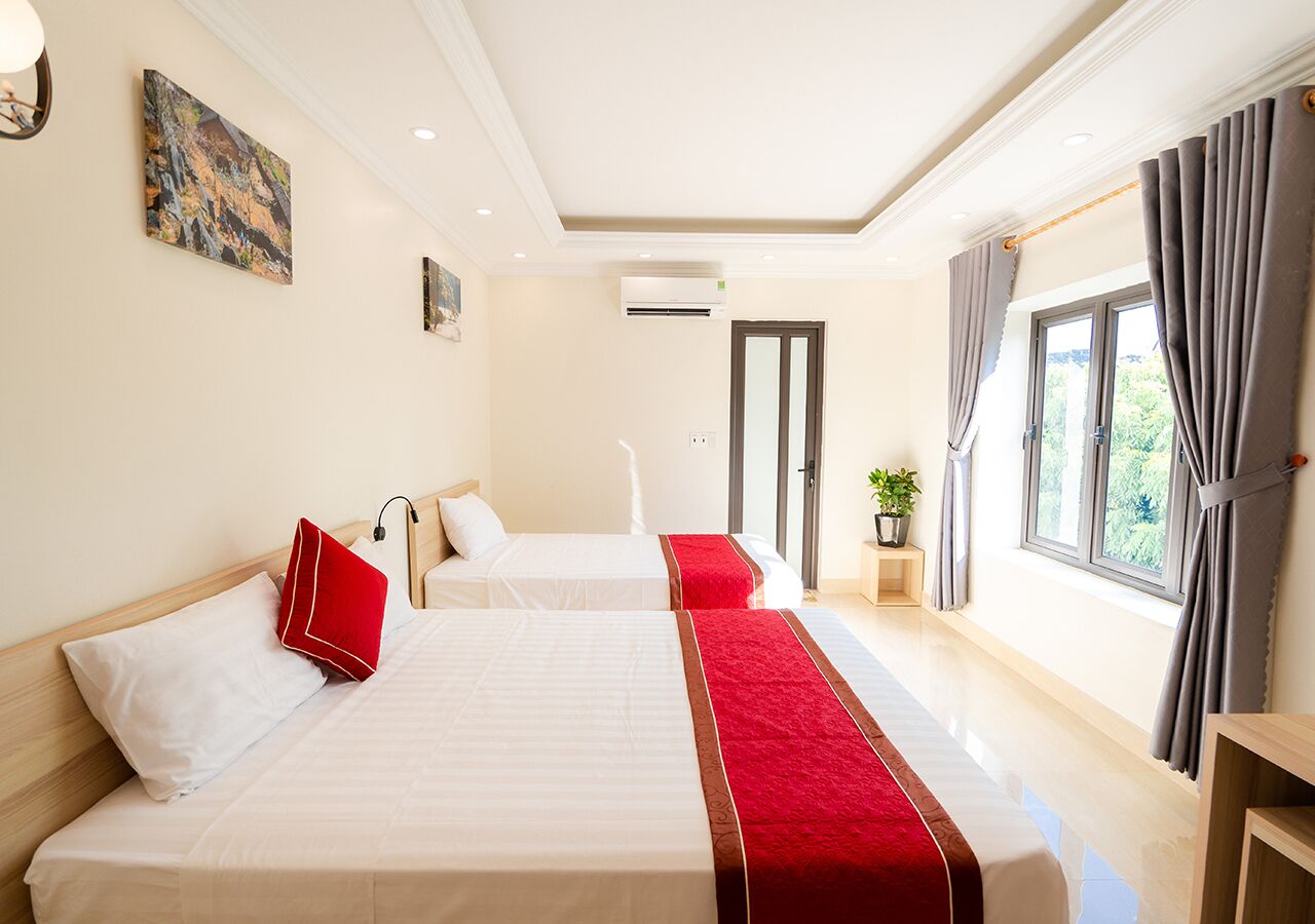 A hotel room with two beds, a red and white bedspread, a potted plant, and a view of greenery through the window.