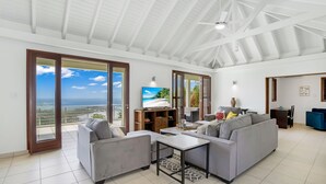 Een ruime woonkamer met een hoge plafond, grote schuifdeuren naar een balkon met uitzicht op zee.