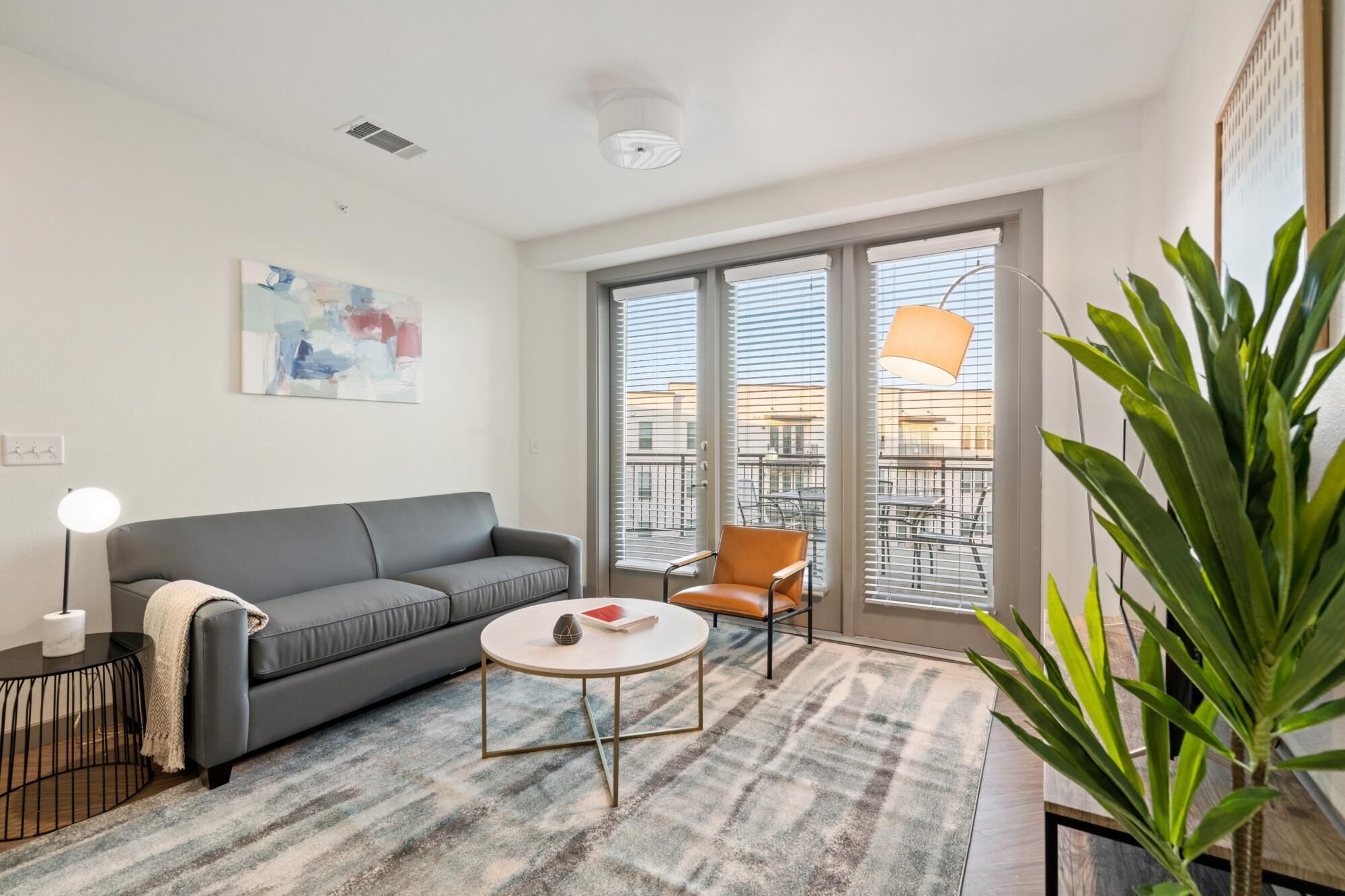 A modern living room with a grey sofa, a round coffee table, and an orange chair. There is a large window with blinds, a ceiling light, and a plant in the corner.