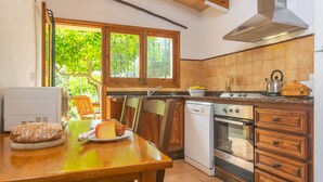 A kitchen with a wooden table, a bread loaf, a plate with a cheese, a window with a view of trees, and a stove. - MARINA (POLLENÇA)  - Charming country house with private swimming pool and free WiFi (Pollença)