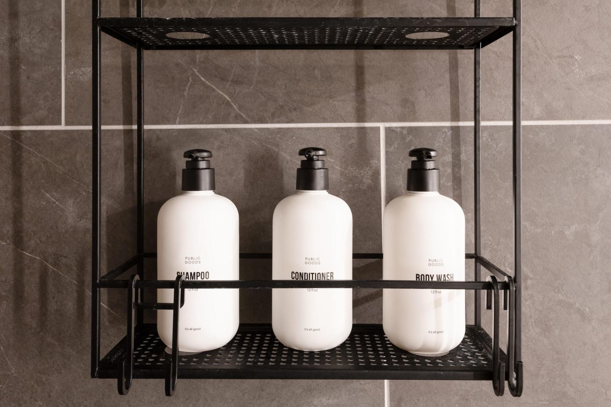 Estante metálico negro con tres botellas blancas etiquetadas como 'Public Goods Shampoo', 'Public Goods Conditioner' y 'Public Goods Body Wash'.