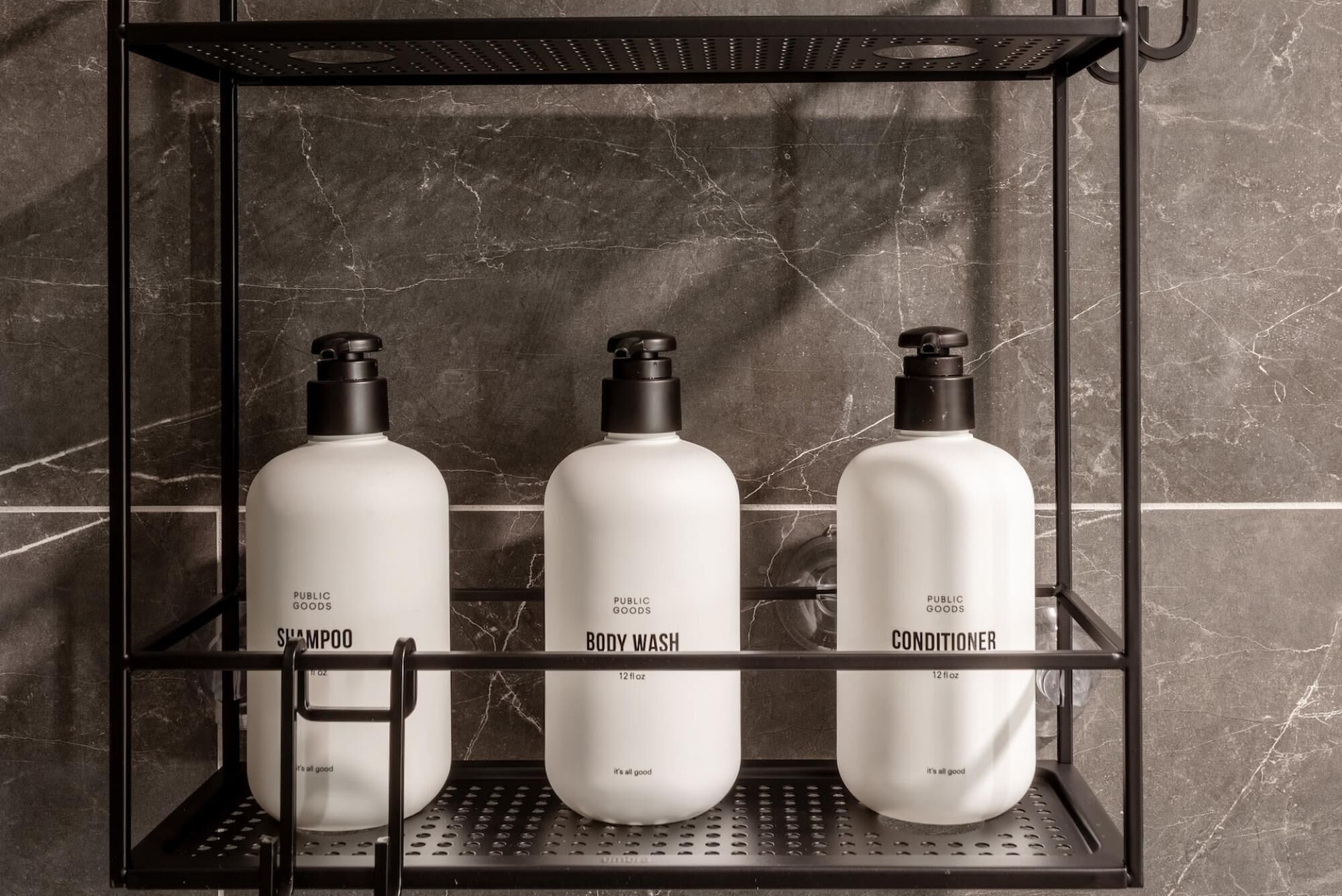 A metal shelf with three white bottles labeled 'PUBLIC GOODS' containing 'SHAMPOO,' 'BODY WASH,' and 'CONDITIONER.'