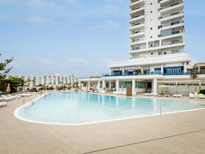 A large swimming pool with sun loungers and a building with balconies.