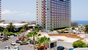 A multi-story hotel with a modern design, featuring a parking area with several cars and palm trees.