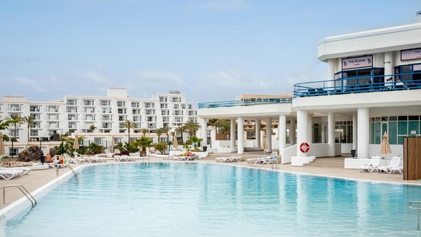 A swimming pool with lounge chairs and umbrellas, surrounded by a hotel with multiple floors and balconies.