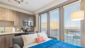 A modern hotel room with a bed, a kitchenette, and large windows with blinds.