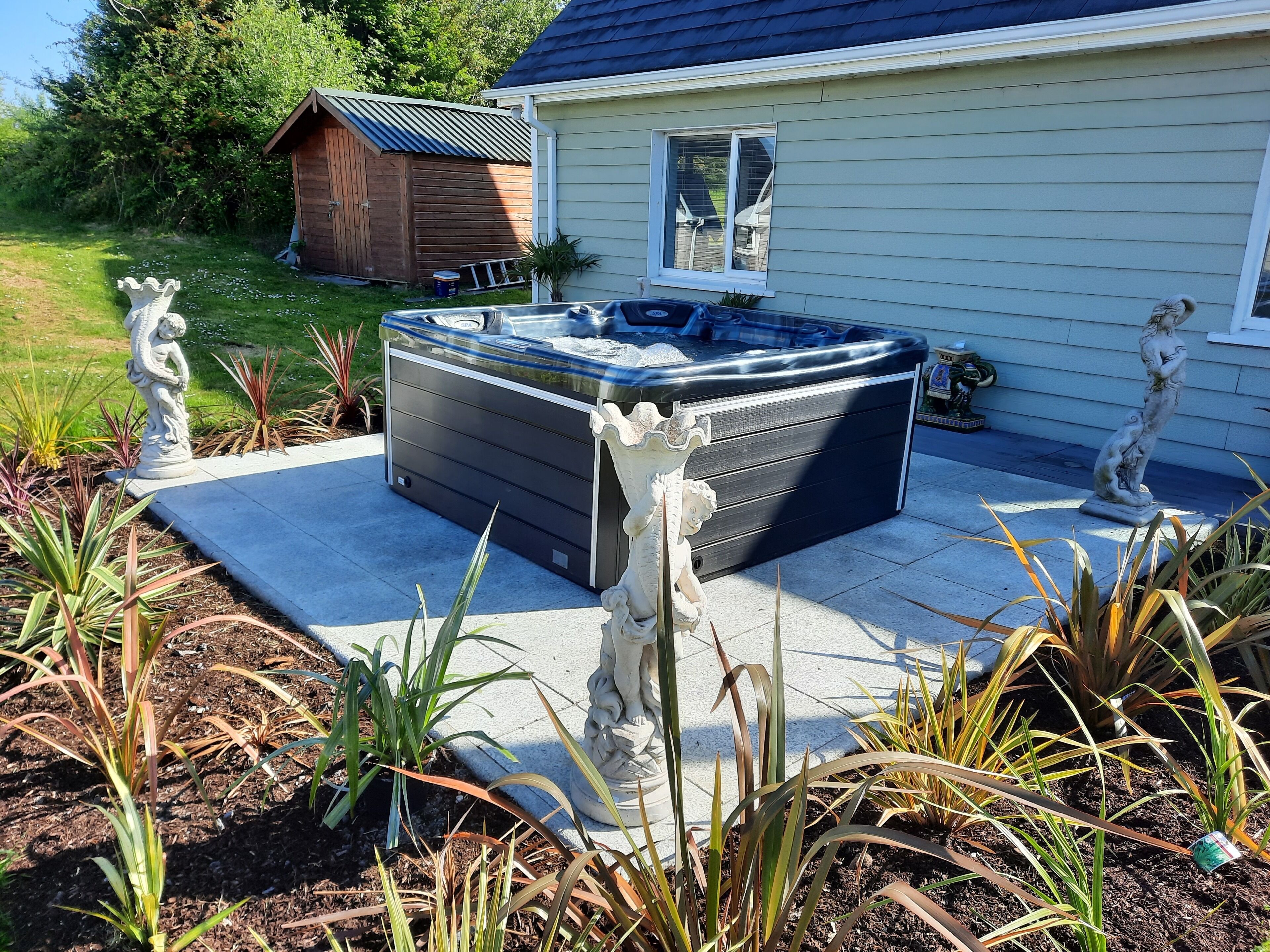 A hot tub in a garden with a wooden shed and a house in the background.