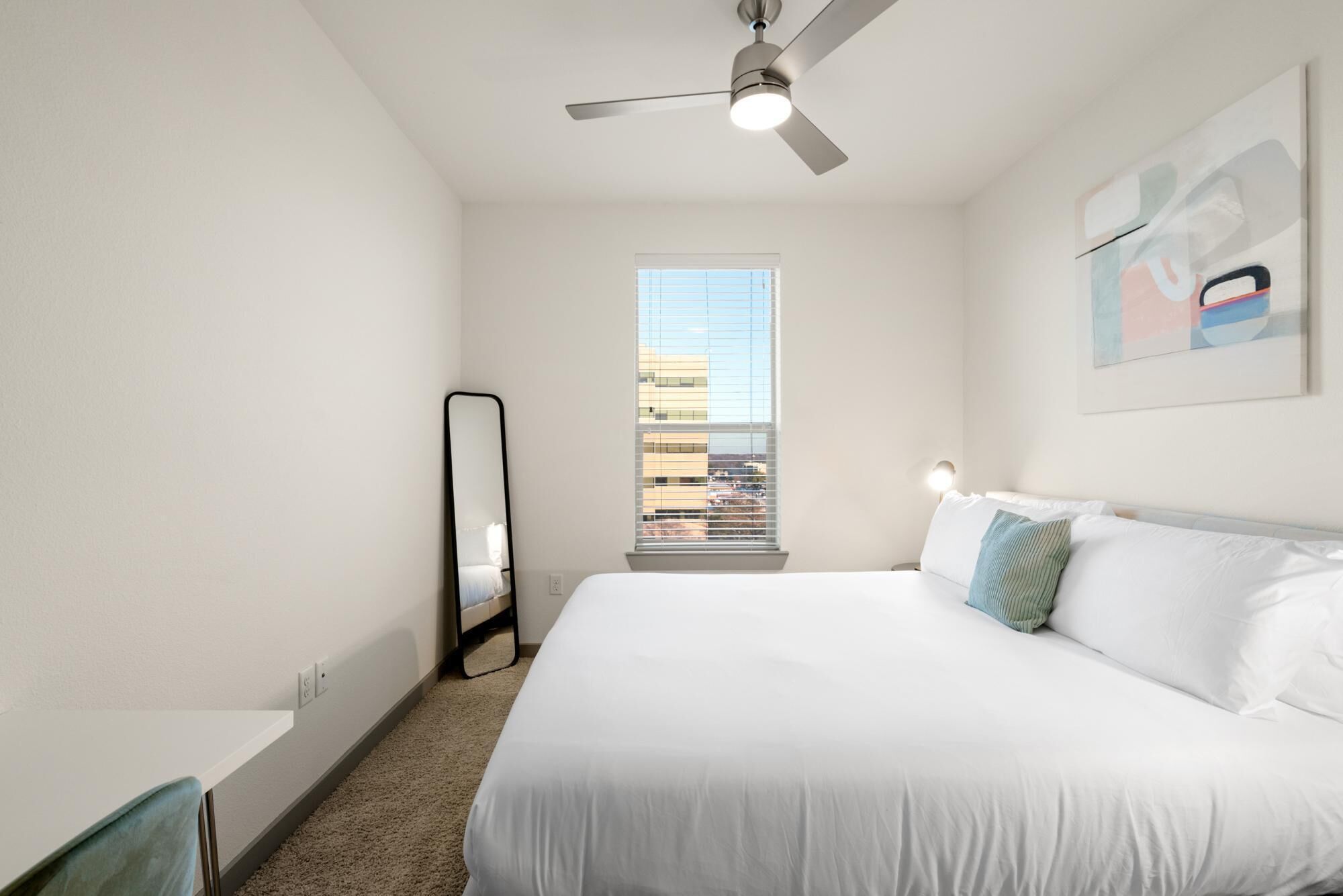 Une chambre d’hôtel avec un grand lit, un ventilateur de plafond, une fenêtre avec des stores, une table de chevet et une œuvre d’art accrochée au mur.