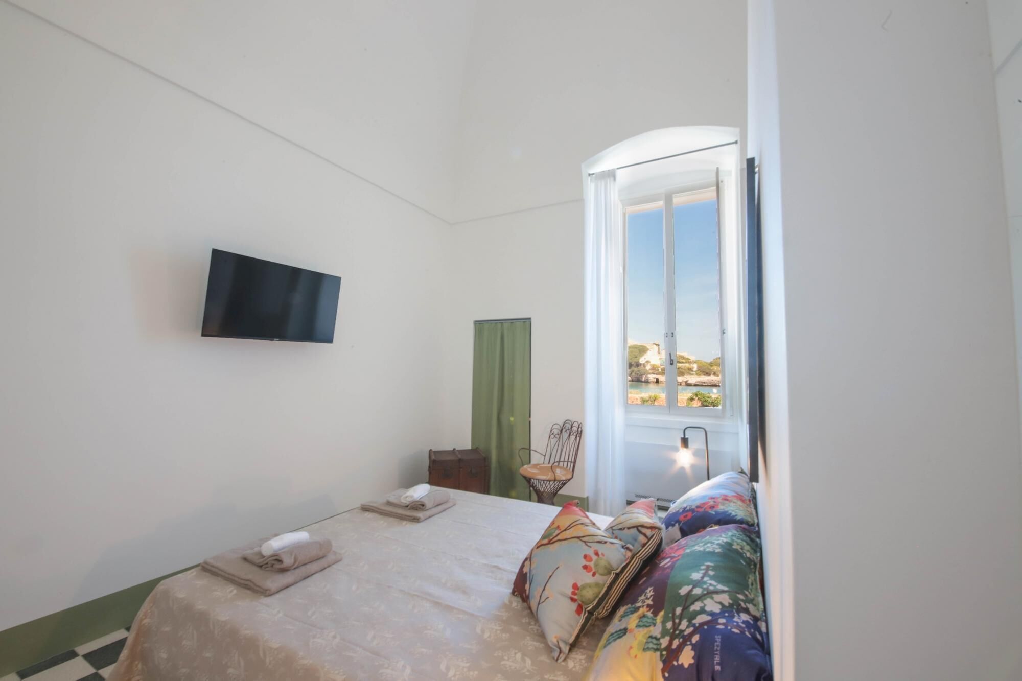 A bedroom with a bed, a flat-screen TV mounted on the wall, a window with a view of a coastal area, and a chair near the window.