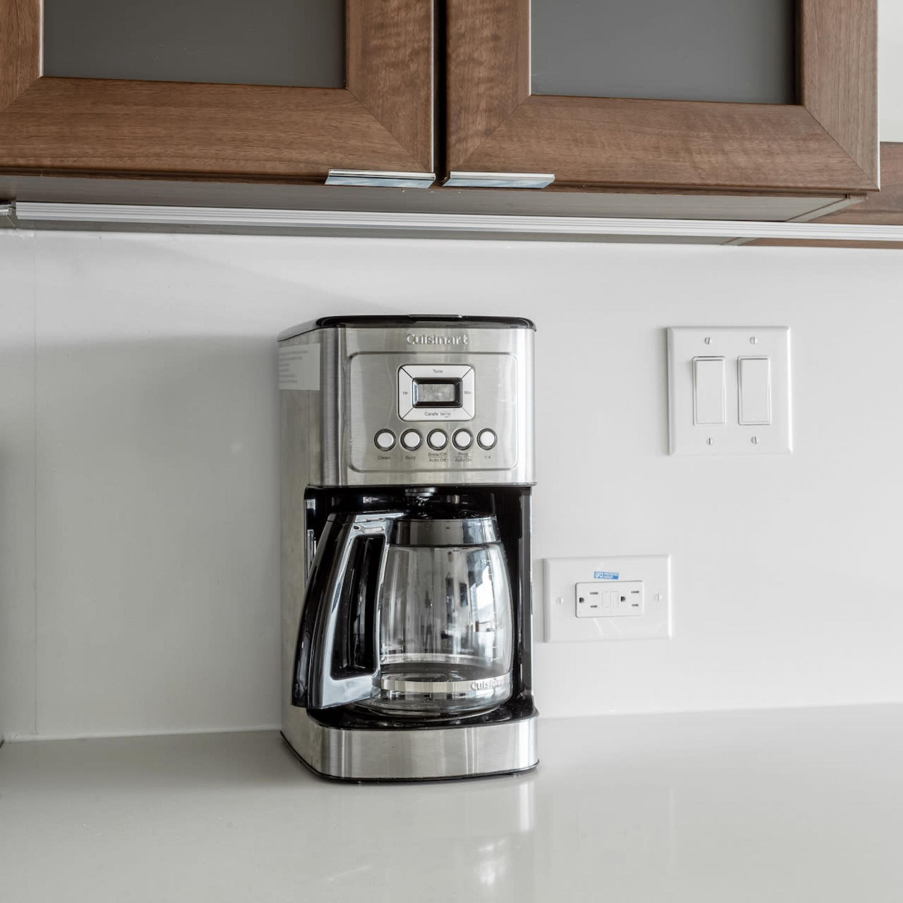 A stainless steel Cuisinart coffee maker on a white countertop with a wooden cabinet above and a wall socket below.