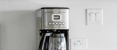 A stainless steel Cuisinart coffee maker on a white countertop with a wooden cabinet above and a wall socket below.