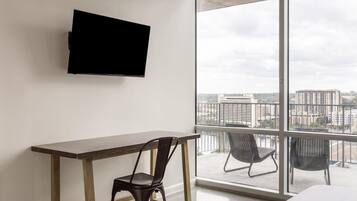 A modern hotel room with a large window, a flat-screen TV mounted on the wall, a wooden desk, and two chairs.