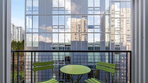 Een balkon met een groene tafel en twee stoelen, een metalen hekwerk en uitzicht op hoge gebouwen door grote ramen.