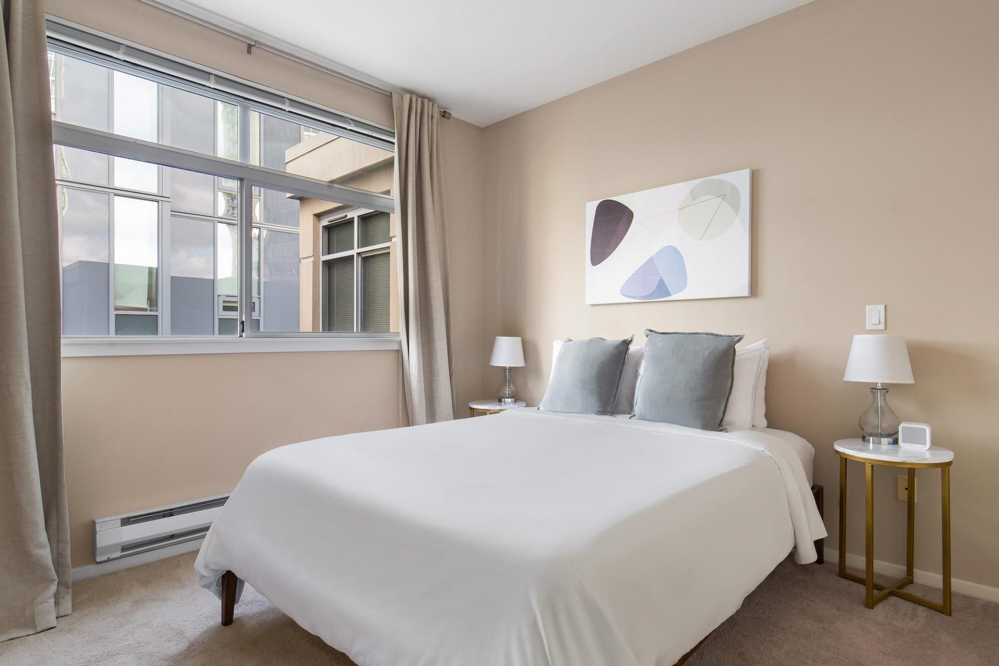 A hotel room with a large bed, two bedside tables with lamps, a window with curtains, and a wall-mounted abstract painting.