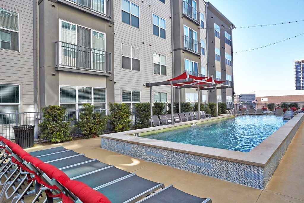 A swimming pool with lounge chairs and a covered seating area, surrounded by a building with multiple balconies and windows.