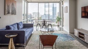 Un salon moderne avec un canapé bleu, une table basse ronde et un fauteuil marron. Une grande fenêtre offre une vue sur la ville.