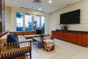 A modern living room with a sofa, ottoman, and coffee table, a TV mounted on the wall, and a large window with a view of buildings.