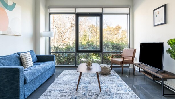 A modern living room with a blue sofa, a coffee table, a TV, and a large window offering a view of trees. - Alexandria Alluring 2BD 2BA Apartment (Alexandria)