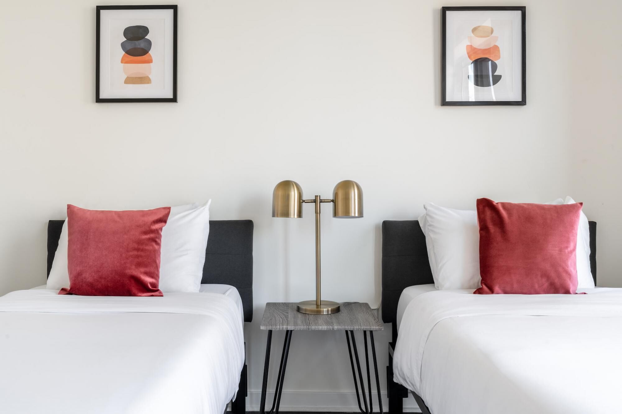 A hotel room with two beds, each with white bedding and red pillows. There is a floor lamp with a gold base and two lampshades. Two framed artworks are hung on the wall above the beds.