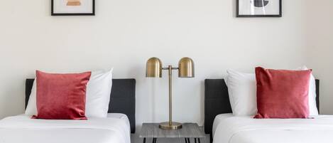 A hotel room with two beds, each with white bedding and red pillows. There is a floor lamp with a gold base and two lampshades. Two framed artworks are hung on the wall above the beds.